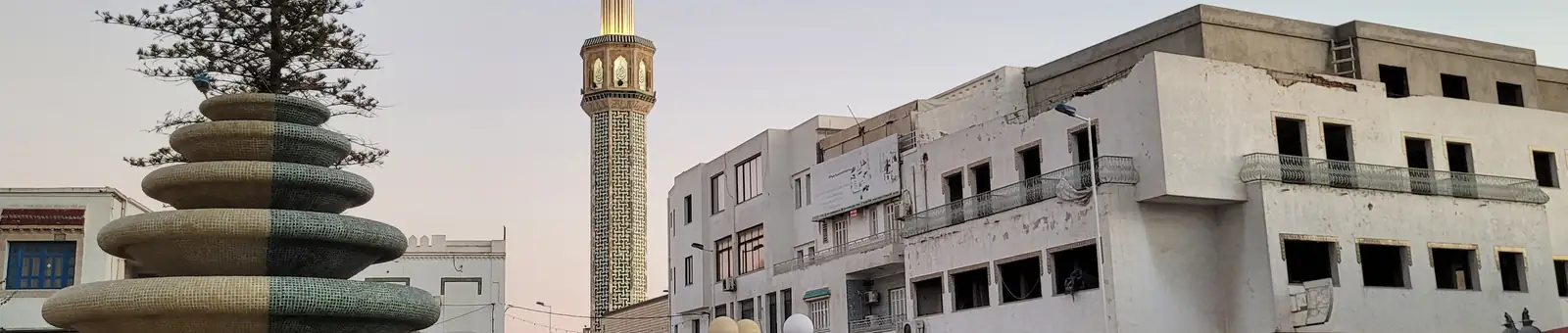 Place de Nabeul à la tombée du jour : une fontaine, un minaret éclairé, des passants et des voitures